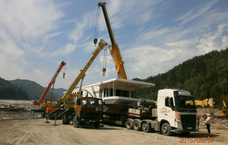 Two cranes are lifting the houseboat over the road train.