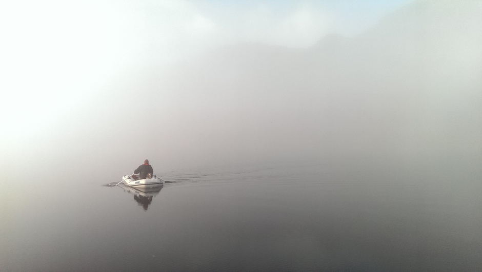 The instructor rows in the mist to the boat after the dog is walking on the shore.